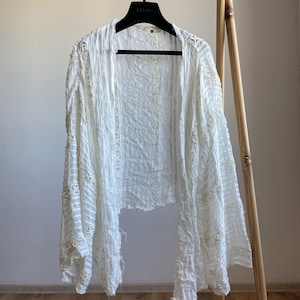 Op de afbeelding: Een witte top met open voorkant en lange mouwen, hangend aan een houten rek. De top heeft een getextureerde stof met delicate kanten details langs de mouwen en randen. Het kledingstuk is lichtgewicht en lijkt gemaakt van een zacht materiaal.