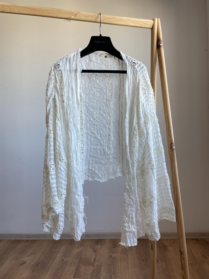 Op de afbeelding: Een witte top met open voorkant en lange mouwen, hangend aan een houten rek. De top heeft een getextureerde stof met delicate kanten details langs de mouwen en randen. Het kledingstuk is lichtgewicht en lijkt gemaakt van een zacht materiaal.