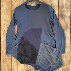 Op de afbeelding: Een tuniek met lange mouwen in tinten donkerblauw en antracietgrijs, voorzien van een patchworkontwerp met gevarieerde texturen, waaronder gladde stof, een fijnmazig paneel, een donker suède-achtig gedeelte en een gewatteerd stoffen paneel.
