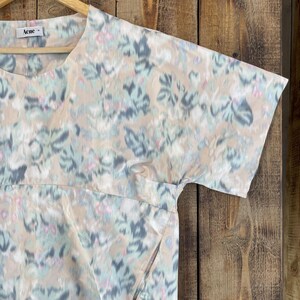 Puede incluir: Una blusa de manga corta con base beige y un patr&oacute;n floral abstracto y difuminado en tonos azul claro, gris oscuro y rosa suave. La prenda presenta un cuello redondo, una costura horizontal en el pecho y una etiqueta de la marca Acne.