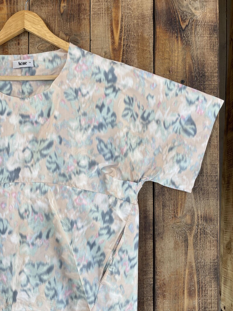 Puede incluir: Una blusa de manga corta con base beige y un patr&oacute;n floral abstracto y difuminado en tonos azul claro, gris oscuro y rosa suave. La prenda presenta un cuello redondo, una costura horizontal en el pecho y una etiqueta de la marca Acne.