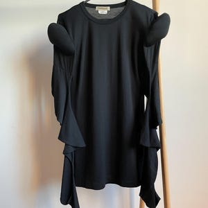 Puede incluir: Camiseta negra de manga larga con mangas con volantes y cuello redondo. La camiseta est&aacute; colgada en una percha de madera.