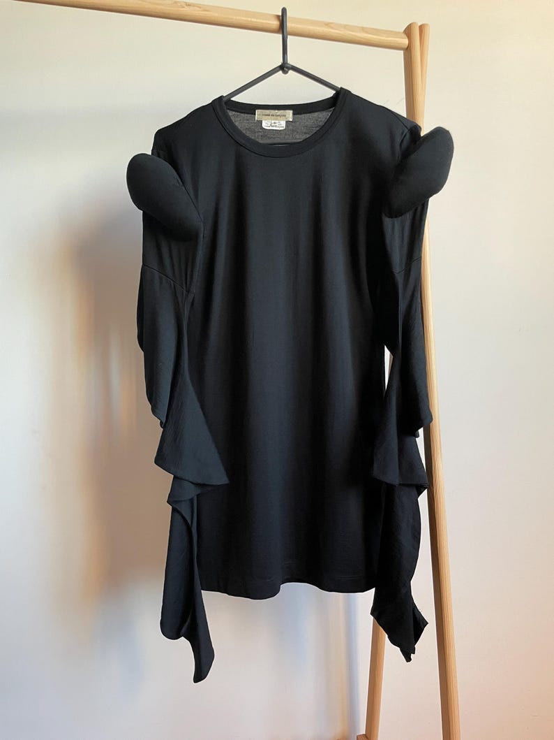 Puede incluir: Camiseta negra de manga larga con mangas con volantes y cuello redondo. La camiseta est&aacute; colgada en una percha de madera.