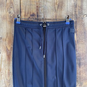 May include: A dark navy blue skirt featuring a full-length central zip, a drawstring waistband with silver-toned eyelets, and vertical stitched pleats. A small rectangular patch with a simple face icon is attached to the upper right side.
