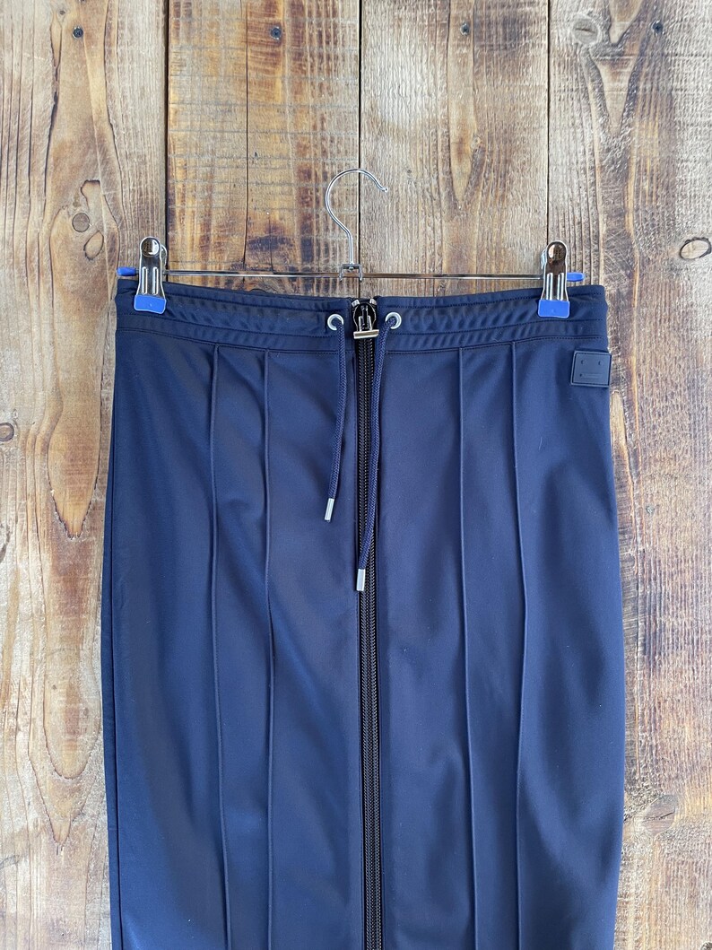 May include: A dark navy blue skirt featuring a full-length central zip, a drawstring waistband with silver-toned eyelets, and vertical stitched pleats. A small rectangular patch with a simple face icon is attached to the upper right side.