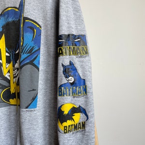 May include: Grey sweatshirt with a large Batman graphic on the front and three smaller Batman graphics on the sleeve. The graphics are in blue, yellow and black.