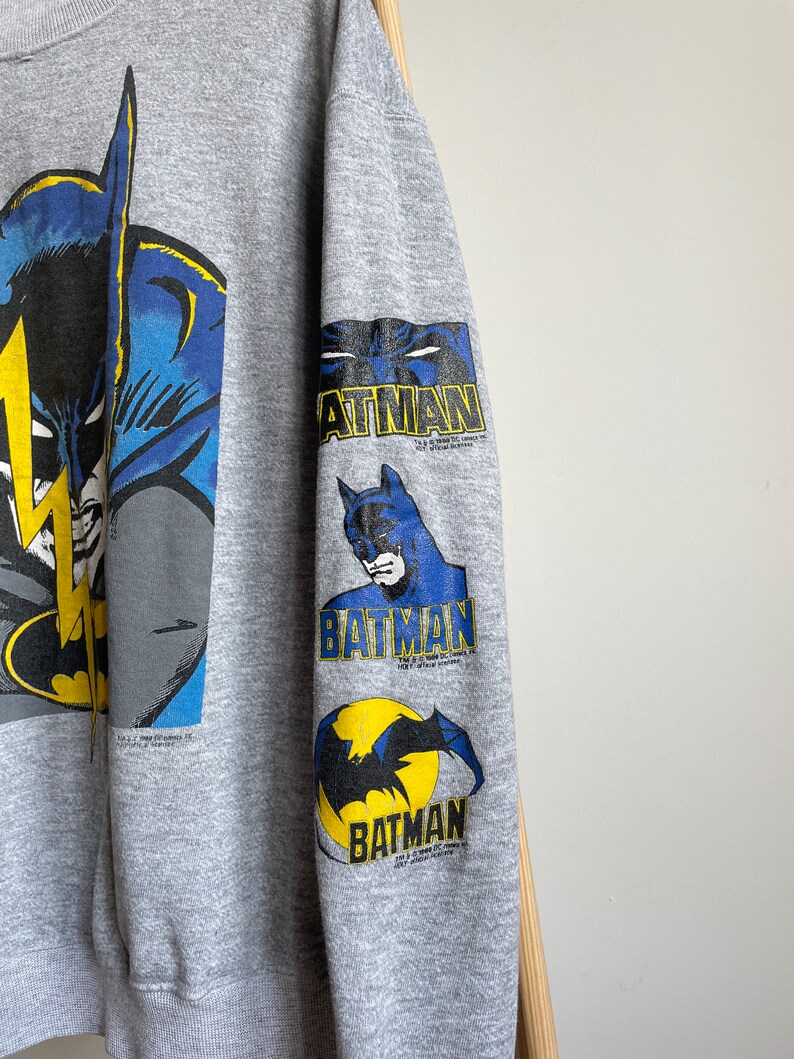 May include: Grey sweatshirt with a large Batman graphic on the front and three smaller Batman graphics on the sleeve. The graphics are in blue, yellow and black.