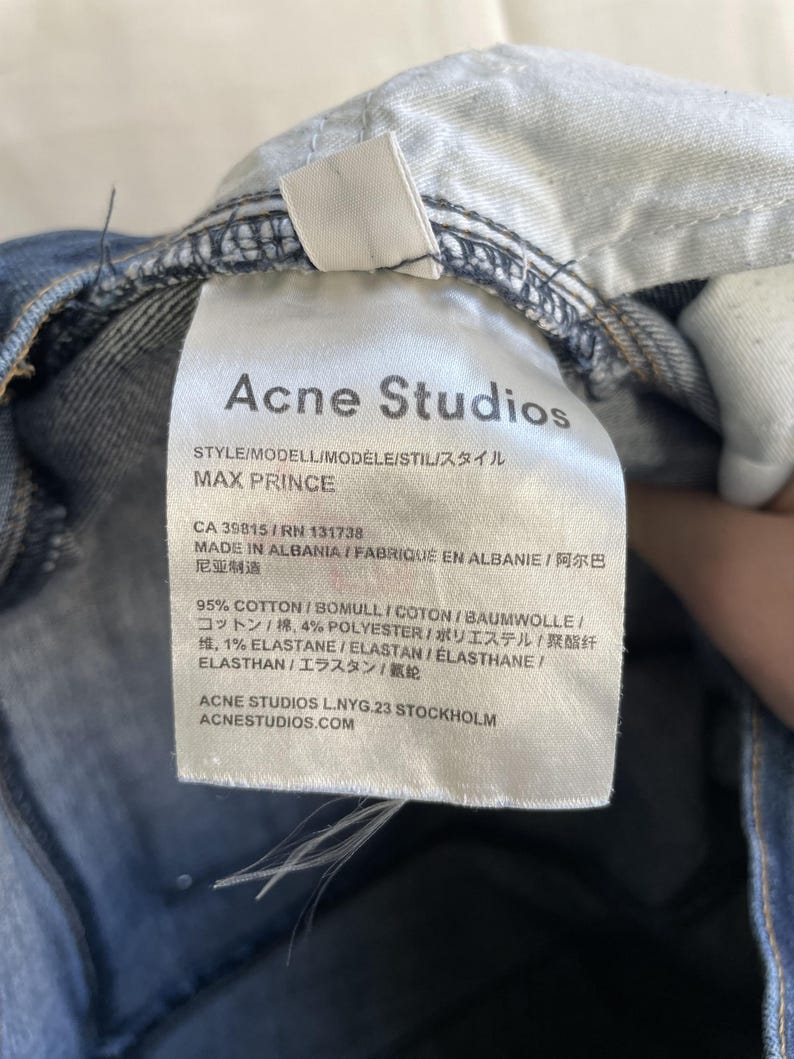 Puede incluir: Primer plano de una etiqueta blanca de vaqueros Acne Studios, con texto negro que detalla el estilo "MAX PRINCE" y la composici&oacute;n de la tela. Los vaqueros son de mezclilla azul.