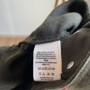 May include: Close-up of a dark green garment with a white tag. The tag includes text in multiple languages, including English, and washing instructions. The garment appears to be made of a cotton and linen blend.