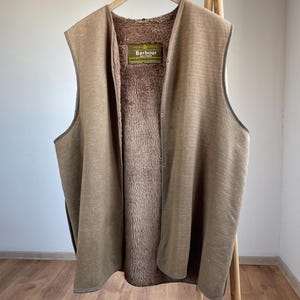 May include: A brown vest with a soft, furry interior and a smooth exterior. The vest is hanging on a wooden hanger. The label reads "Barbour".