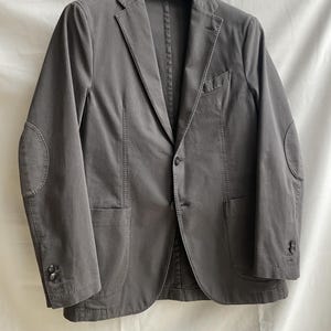 May include: A gray blazer with elbow patches, two front pockets, and a single-button closure. The jacket is hanging on a black hanger against a white background. The blazer has buttons on the sleeves.