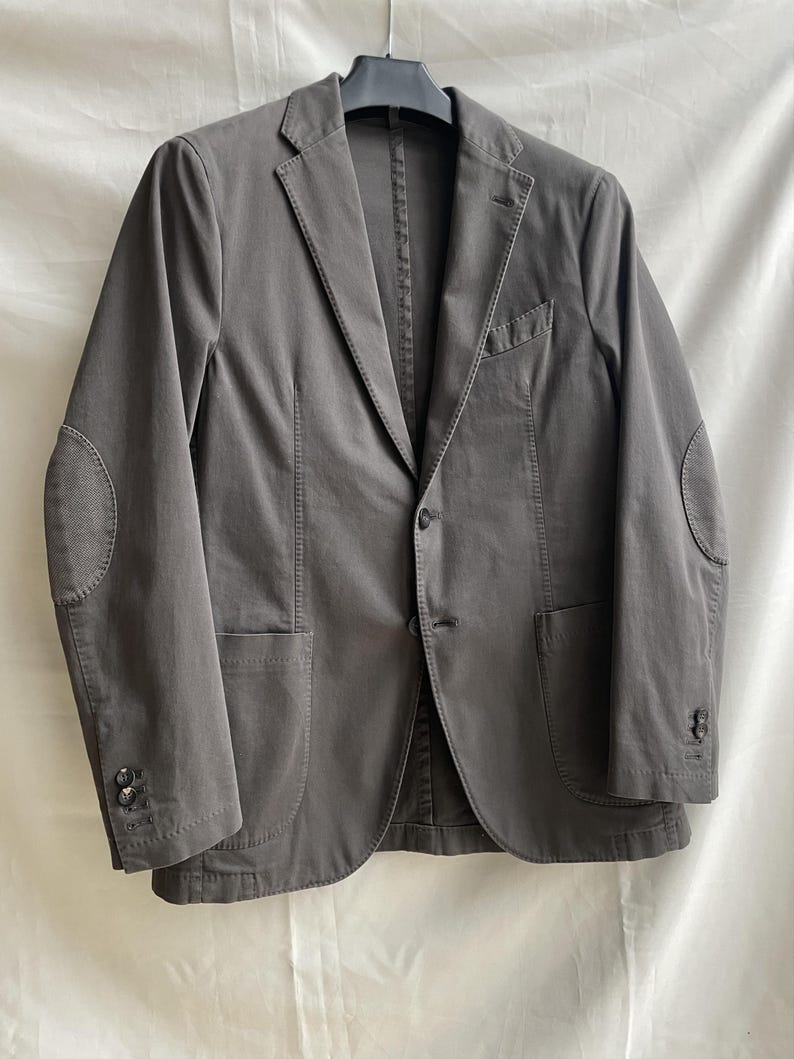 May include: A gray blazer with elbow patches, two front pockets, and a single-button closure. The jacket is hanging on a black hanger against a white background. The blazer has buttons on the sleeves.