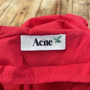 May include: A bright red garment featuring a white rectangular fabric label stitched onto the waistband. The label displays the black text Acne and the number 40 in a smaller font, with a green mark visible next to the size number.