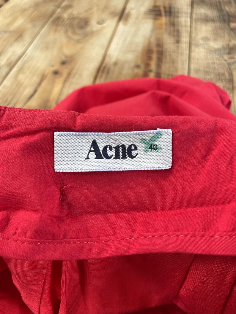 May include: A bright red garment featuring a white rectangular fabric label stitched onto the waistband. The label displays the black text Acne and the number 40 in a smaller font, with a green mark visible next to the size number.