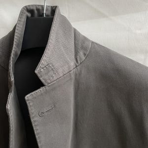 May include: Close-up of a gray jacket with a textured collar. The jacket has a single button closure and is hanging on a black hanger. The fabric appears to be a medium-weight material.