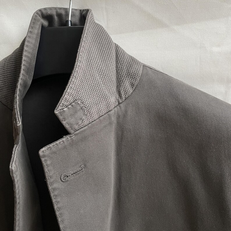 May include: Close-up of a gray jacket with a textured collar. The jacket has a single button closure and is hanging on a black hanger. The fabric appears to be a medium-weight material.