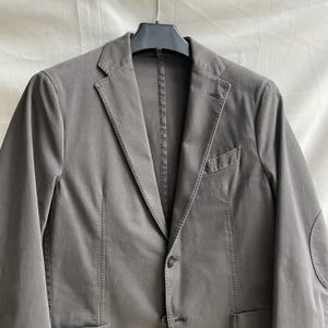 May include: A gray blazer with a two-button closure, notched lapels, and a chest pocket. The jacket has elbow patches and is hanging on a black hanger. The fabric appears to be a cotton blend.