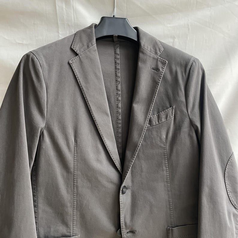 May include: A gray blazer with a two-button closure, notched lapels, and a chest pocket. The jacket has elbow patches and is hanging on a black hanger. The fabric appears to be a cotton blend.