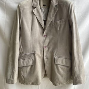 May include: A light beige suede blazer with a notched lapel, two front pockets, and a single-breasted button closure. The jacket has a tailored fit and is hanging on a black hanger against a white background.
