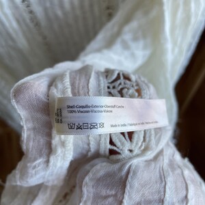 Op de afbeelding: Close-up van witte stof met een label. Het label bevat tekst in meerdere talen, waaronder "Shell-Coquille-Exterior-Oberstoff-Conche" en "100% Viscose-Viscosa-Viskose". De stof heeft een delicaat, open weefpatroon.