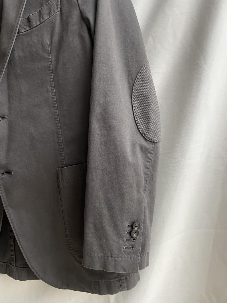 May include: A charcoal gray blazer with a notched lapel, a single-breasted closure, and a patch pocket. The blazer features elbow patches and buttoned cuffs. The fabric appears to be a medium-weight cotton or blend.