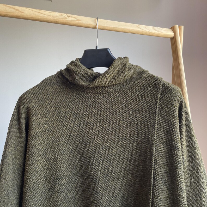 Peut inclure: Pull &agrave; col roul&eacute; vert olive suspendu &agrave; un portant en bois. Le pull pr&eacute;sente un motif en tricot textur&eacute; et un col haut et repli&eacute;. Les manches sont longues et le style est d&eacute;contract&eacute; et confortable.