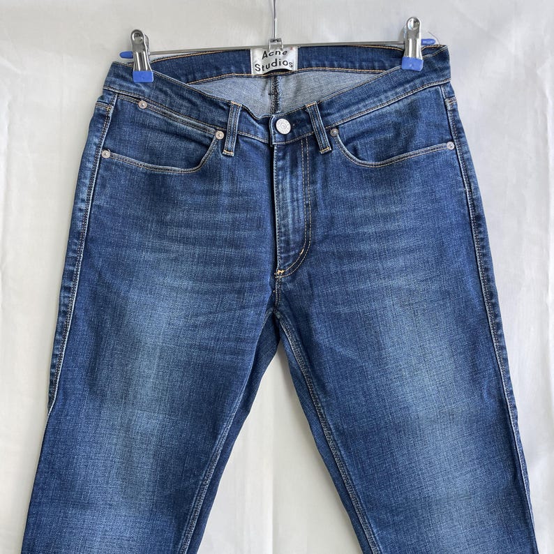 Puede incluir: Vaqueros de mezclilla azules con un dise&ntilde;o cl&aacute;sico de cinco bolsillos. Los vaqueros tienen cierre de bot&oacute;n y cremallera, trabillas para el cintur&oacute;n y la etiqueta "Acne Studios". La mezclilla tiene un lavado medio con decoloraci&oacute;n sutil y pernera recta.
