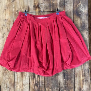 May include: A bright red bubble skirt featuring a high-waisted design with deep vertical pleats and two matching red circular buttons on the left side. The skirt has a voluminous, rounded silhouette that gathers at the bottom hemline.