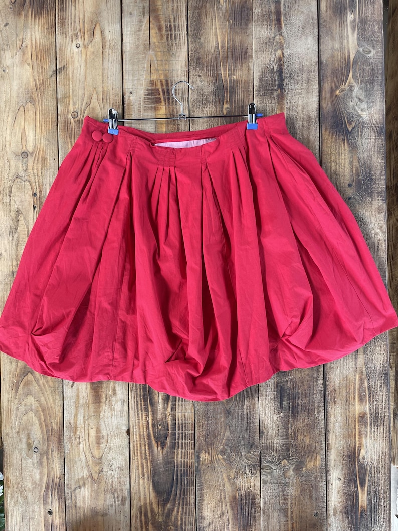 May include: A bright red bubble skirt featuring a high-waisted design with deep vertical pleats and two matching red circular buttons on the left side. The skirt has a voluminous, rounded silhouette that gathers at the bottom hemline.
