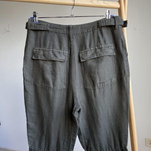 May include: Dark olive green pants with two back pockets and a belt. The pants are made of a soft, textured fabric and hang from a wooden clothing rack. The pants have a relaxed fit and a casual style.