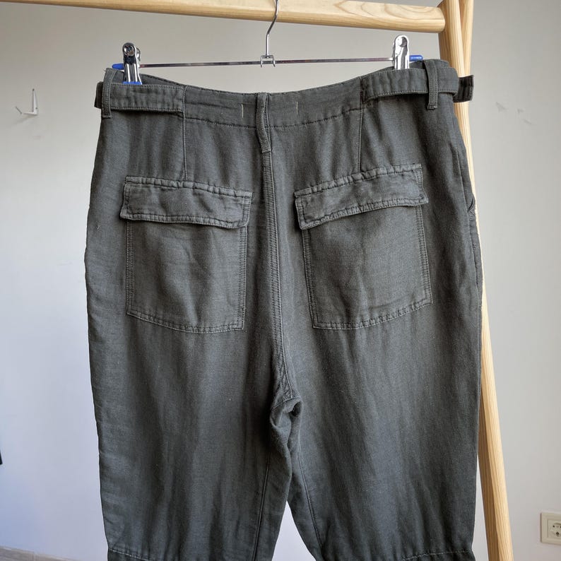 May include: Dark olive green pants with two back pockets and a belt. The pants are made of a soft, textured fabric and hang from a wooden clothing rack. The pants have a relaxed fit and a casual style.