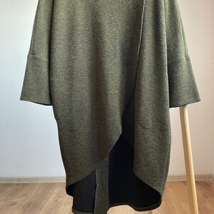 Peut inclure: Cardigan en tricot vert olive avec des manches larges et un ourlet asym&eacute;trique. Le pull &agrave; manches longues pr&eacute;sente un motif en tricot textur&eacute; et une coupe d&eacute;contract&eacute;e. Le v&ecirc;tement est pr&eacute;sent&eacute; sur un portant en bois. La longueur du cardigan est d'environ 90 cm.