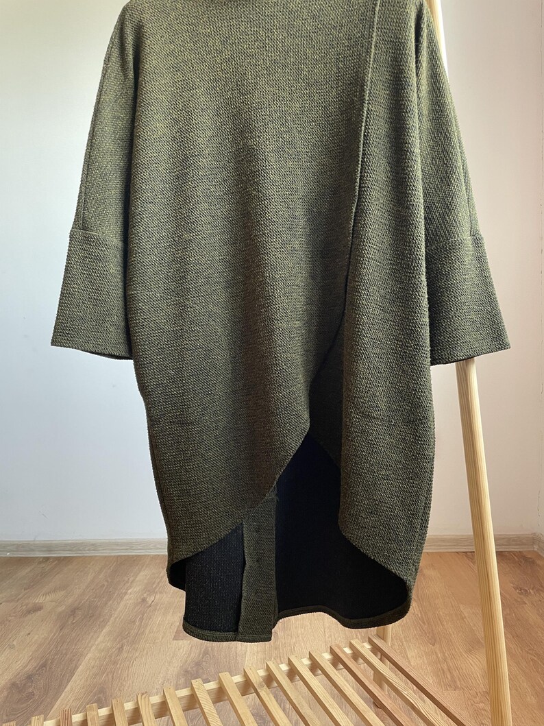 Peut inclure: Cardigan en tricot vert olive avec des manches larges et un ourlet asym&eacute;trique. Le pull &agrave; manches longues pr&eacute;sente un motif en tricot textur&eacute; et une coupe d&eacute;contract&eacute;e. Le v&ecirc;tement est pr&eacute;sent&eacute; sur un portant en bois. La longueur du cardigan est d'environ 90 cm.