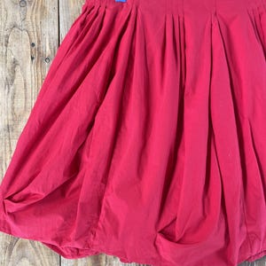 May include: A bright red bubble skirt featuring a high-waisted design with vertical pleats along the waistband. The skirt has a voluminous, rounded silhouette created by the gathered hemline, which tucks inward to form a distinct bubble shape.