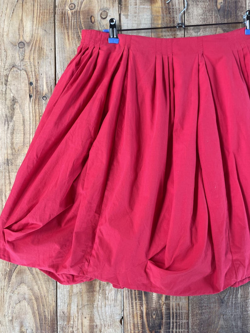 May include: A bright red bubble skirt featuring a high-waisted design with vertical pleats along the waistband. The skirt has a voluminous, rounded silhouette created by the gathered hemline, which tucks inward to form a distinct bubble shape.