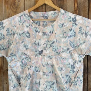 Puede incluir: Una blusa de manga corta con cuello redondo y color base beige que presenta un patr&oacute;n floral abstracto y difuminado en tonos azul claro, gris y rosa suave. La prenda incluye una costura horizontal en el pecho y la etiqueta Acne.