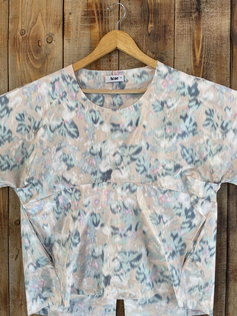 Puede incluir: Una blusa de manga corta con cuello redondo y color base beige que presenta un patr&oacute;n floral abstracto y difuminado en tonos azul claro, gris y rosa suave. La prenda incluye una costura horizontal en el pecho y la etiqueta Acne.
