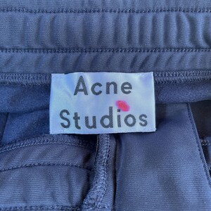 May include: A dark navy blue garment featuring a white rectangular fabric label stitched at the waistband. The label displays the text Acne Studios in black font with a small pink dot mark located above the letter i in the word Studios.