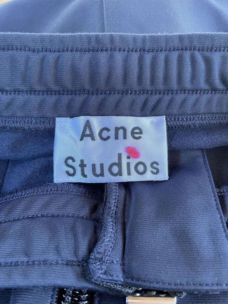 May include: A dark navy blue garment featuring a white rectangular fabric label stitched at the waistband. The label displays the text Acne Studios in black font with a small pink dot mark located above the letter i in the word Studios.