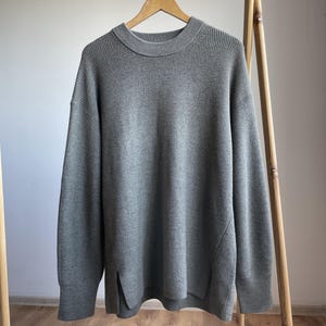 Puede incluir: Un suéter gris con textura acanalada cuelga de una percha de madera. El suéter tiene cuello redondo, mangas largas y aberturas laterales. La prenda se exhibe en un perchero de madera.