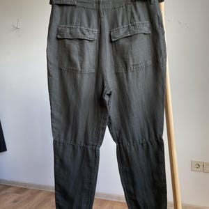 May include: Dark olive green pants with two back pockets and a belt. The pants are hanging on a wooden rack. The fabric appears to be a linen blend. The pants have a relaxed fit and a casual style.