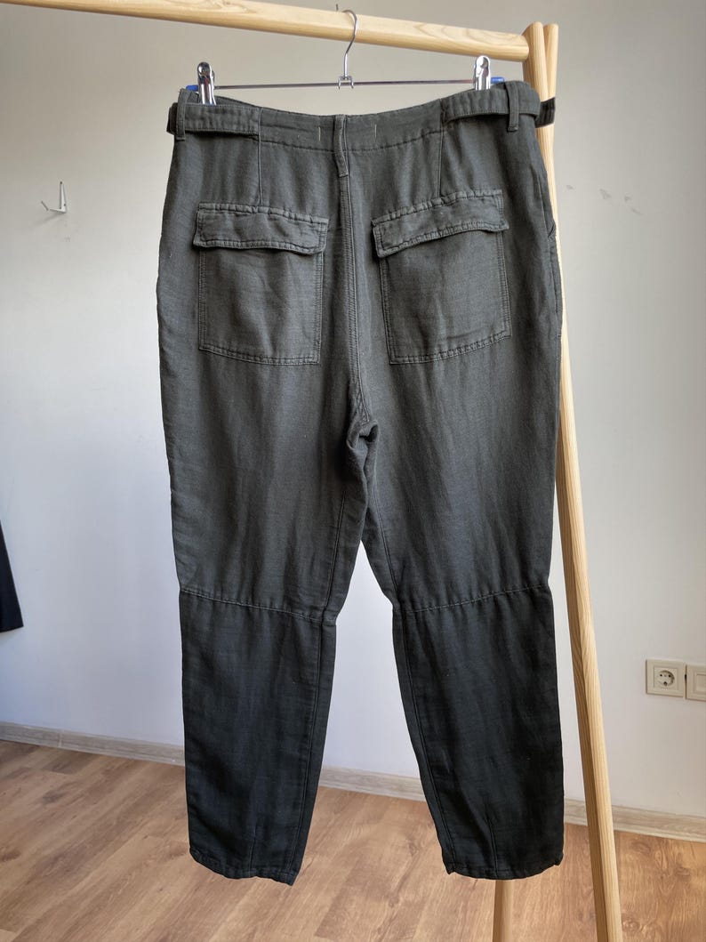 May include: Dark olive green pants with two back pockets and a belt. The pants are hanging on a wooden rack. The fabric appears to be a linen blend. The pants have a relaxed fit and a casual style.