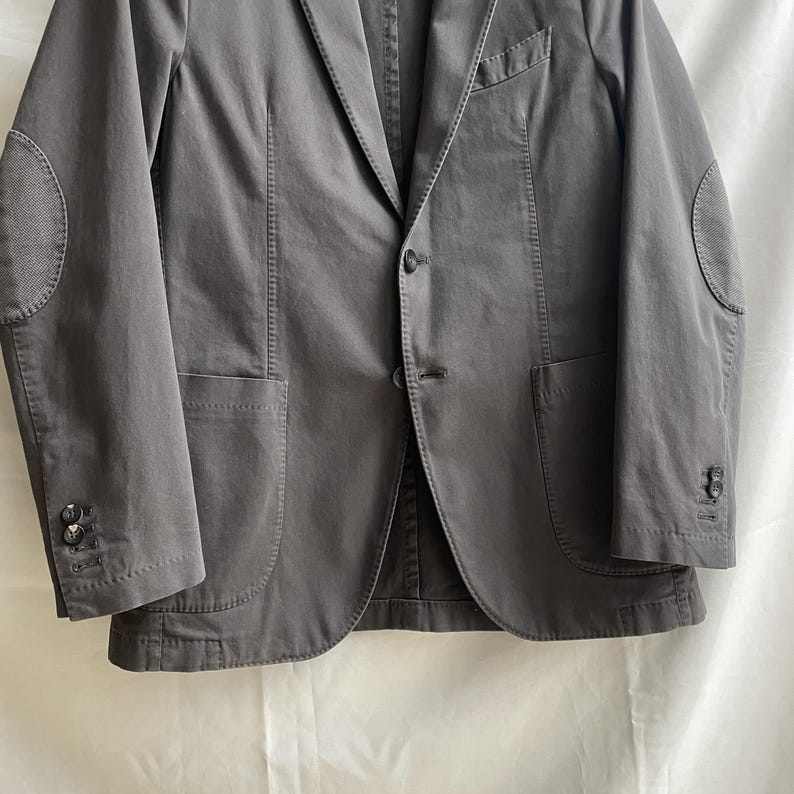 May include: A gray blazer with elbow patches. The jacket has a notched lapel, two front pockets, and buttoned cuffs. The elbow patches are a lighter gray, textured fabric. The blazer is buttoned at the center and has a tailored fit.