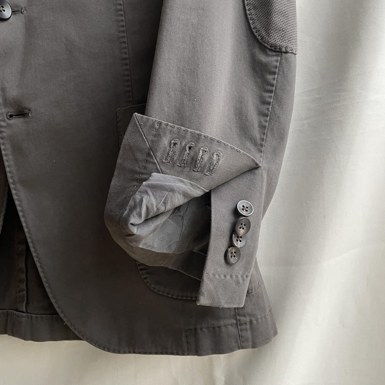 May include: Close-up of a gray blazer with a notched lapel and three buttons on the sleeve cuff. The blazer has a textured elbow patch and a tailored fit. The fabric appears to be a cotton blend.