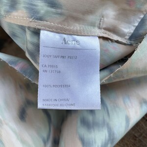 Puede incluir: Una etiqueta de cuidado blanca unida a una tela con un estampado abstracto borroso y multicolor. La etiqueta presenta el texto Acne, JODY TAFF PRT PSS12, CA 39815, RN 131738, 100% POLYESTER y MADE IN CHINA/FABRIQUE AU CHINE en tinta negra.