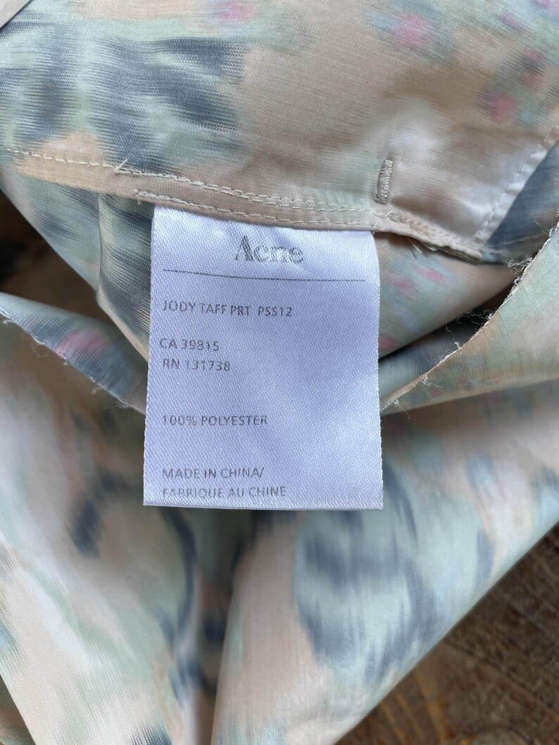 Puede incluir: Una etiqueta de cuidado blanca unida a una tela con un estampado abstracto borroso y multicolor. La etiqueta presenta el texto Acne, JODY TAFF PRT PSS12, CA 39815, RN 131738, 100% POLYESTER y MADE IN CHINA/FABRIQUE AU CHINE en tinta negra.