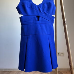 Puede incluir: Un vestido azul vibrante con escote en V y detalles recortados en la cintura. El vestido presenta un corpi&ntilde;o ajustado y una falda acampanada, colgado de una percha de madera. La tela parece lisa y tiene un ligero brillo.