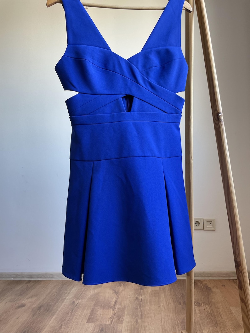 Puede incluir: Un vestido azul vibrante con escote en V y detalles recortados en la cintura. El vestido presenta un corpi&ntilde;o ajustado y una falda acampanada, colgado de una percha de madera. La tela parece lisa y tiene un ligero brillo.