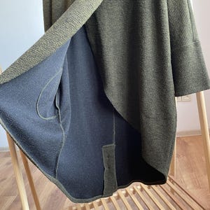 Peut inclure: Cardigan en tricot vert olive avec un int&eacute;rieur bleu marine fonc&eacute; contrastant. Le cardigan pr&eacute;sente un design drap&eacute; unique avec un tissu en tricot textur&eacute;. Les manches sont aux trois quarts et le v&ecirc;tement est pr&eacute;sent&eacute; sur un portant en bois.