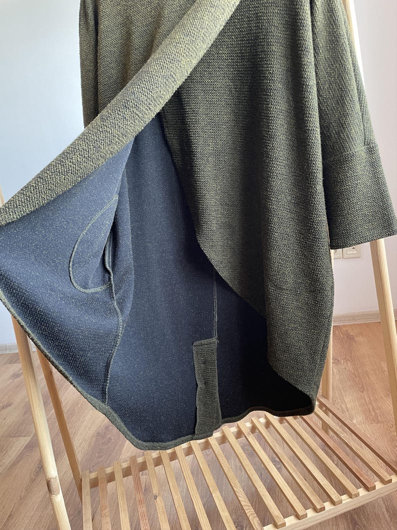 Peut inclure: Cardigan en tricot vert olive avec un int&eacute;rieur bleu marine fonc&eacute; contrastant. Le cardigan pr&eacute;sente un design drap&eacute; unique avec un tissu en tricot textur&eacute;. Les manches sont aux trois quarts et le v&ecirc;tement est pr&eacute;sent&eacute; sur un portant en bois.
